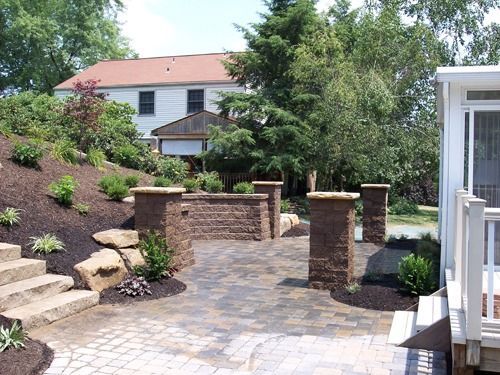 Paved patio with pillars, a retaining wall, and steps. A two-story house is in the background. Landscaping includes trees and shrubs.