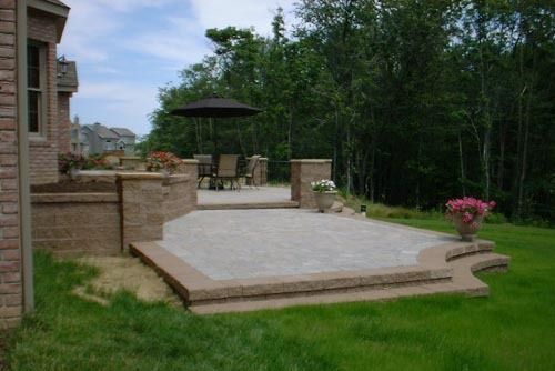 Stone patio with steps and retaining walls, table and chairs with umbrella, next to grassy yard and trees.