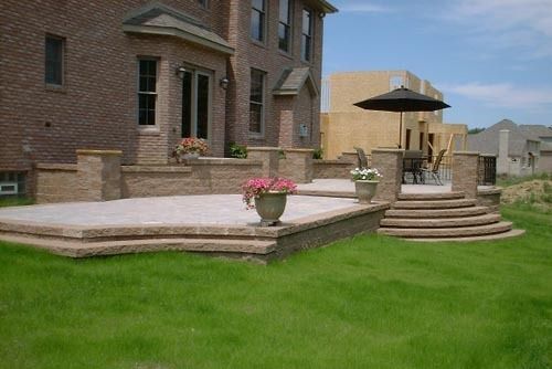 Backyard patio with stone steps, flower pots, and umbrella, in front of a brick house.
