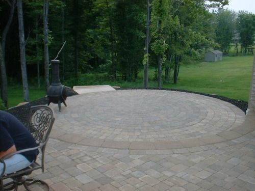 Circular paved patio with fire pit, surrounded by trees and grass, with a seated person on the left.