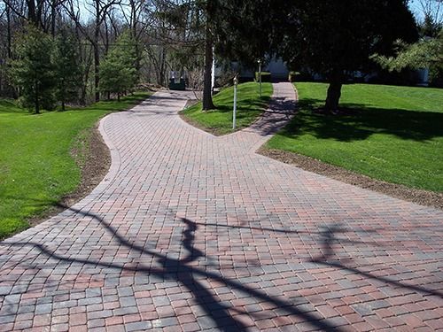 Brick driveway splits, winding through green lawns, with tree shadows.