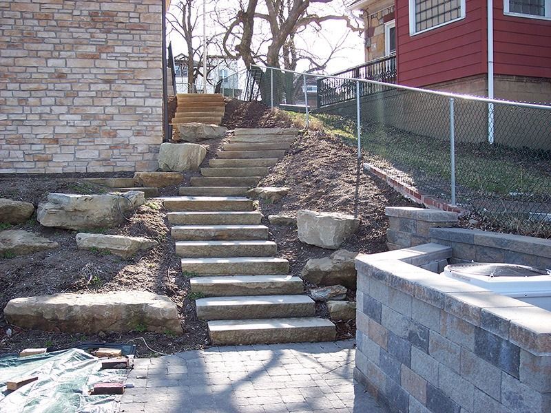 Stone steps lead uphill in a backyard with landscaping; chain link fence and brick wall.