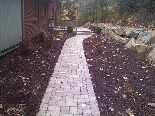Brick pathway winding through a landscaped yard with mulch and rock wall.