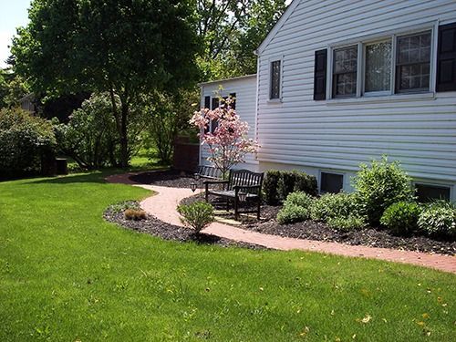 A brick pathway curves through a green lawn towards a white house with black shutters, a bench, and a flowering tree.