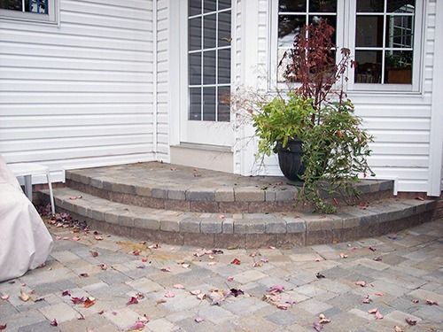 Brick steps leading to a white door, with potted plants on the landing and patio pavers below.