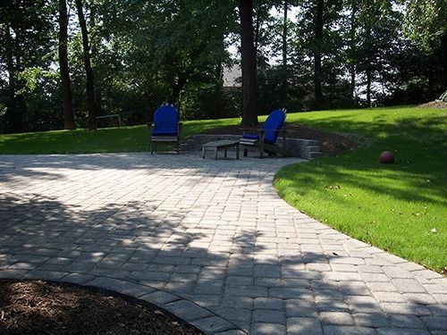 Brick patio with two blue chairs near a fire pit, surrounded by grass and trees.