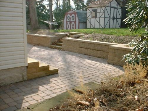 Stone retaining walls flank a paved pathway and steps leading towards a shed and house.