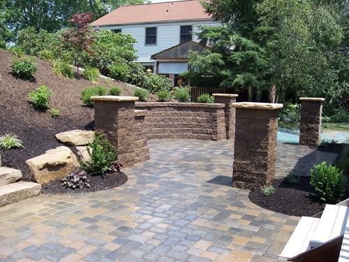 Brick patio with retaining wall, pillars, and landscaping in front of a house.