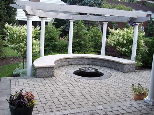Stone patio with a circular bench and pergola, surrounding a small water feature.
