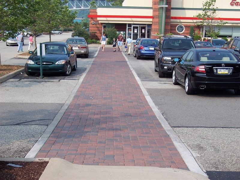 Brick walkway in parking lot, cars parked on either side, people walking towards a building entrance.