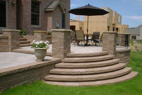 Outdoor patio with curved steps, columns, seating area with umbrella, and a brick house.