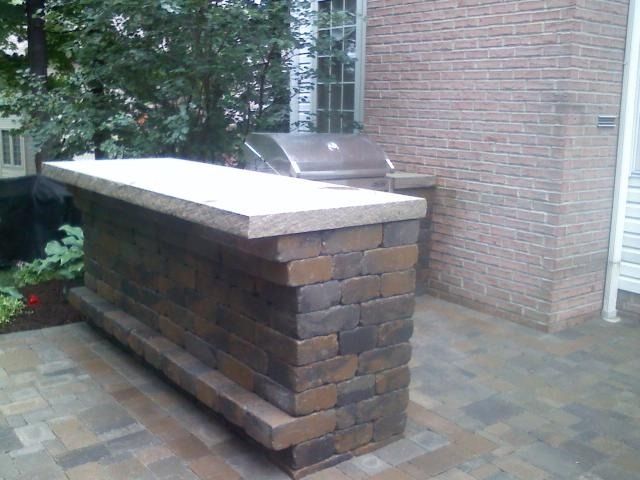 Outdoor brick grilling station with a stone countertop and built-in grill next to a brick building.