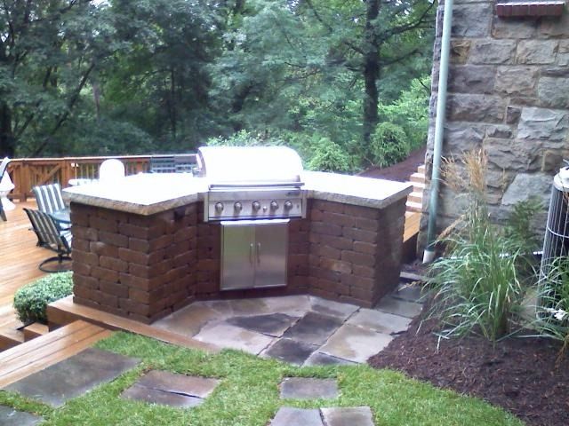 Built-in brick outdoor grill with stainless steel features and stone countertops, near a deck and landscaped yard.