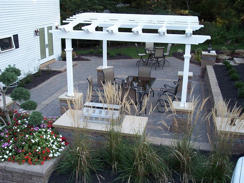 White pergola over patio with seating; low walls with plants and house in background.