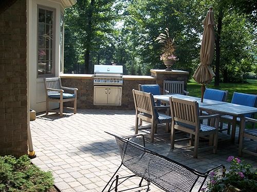 Patio with built-in grill, dining table, chairs, and umbrella; surrounded by trees and a brick wall.
