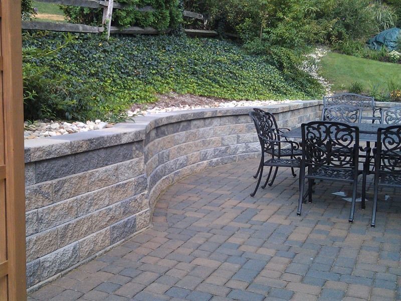 Stone retaining wall curves around a brick patio with wrought iron furniture, hillside in background.