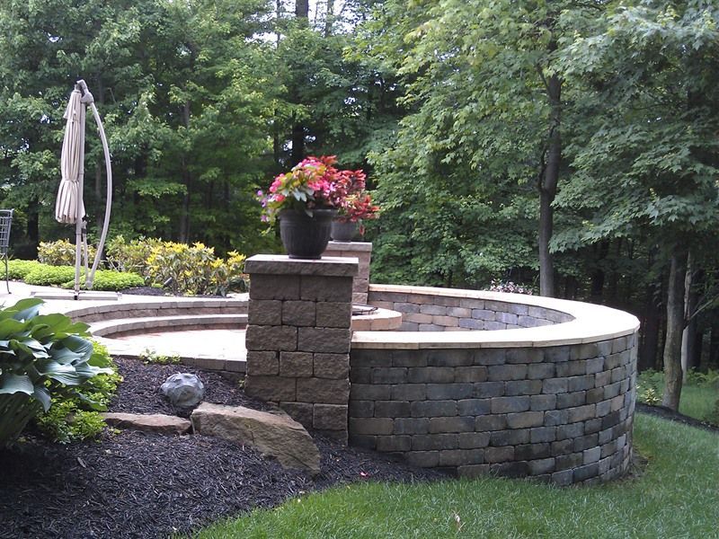 Stone patio with tiered seating, curved wall, flowers in a pot, surrounded by trees and black mulch.