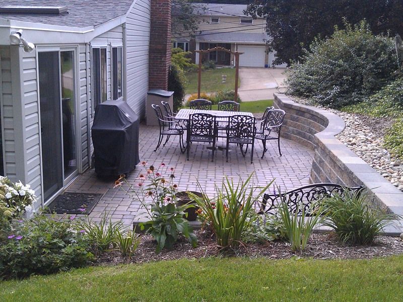 Brick patio with outdoor dining set; landscaping with retaining wall and grassy area.