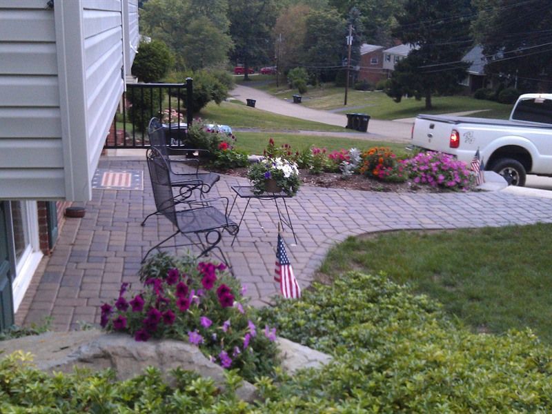 Patio with brick pavers, metal chairs, and flowerbeds near a house and street. A white truck is parked nearby.