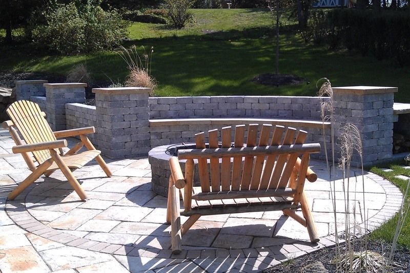 Wooden chairs on a stone patio with a built-in stone bench and fire pit in a backyard setting.