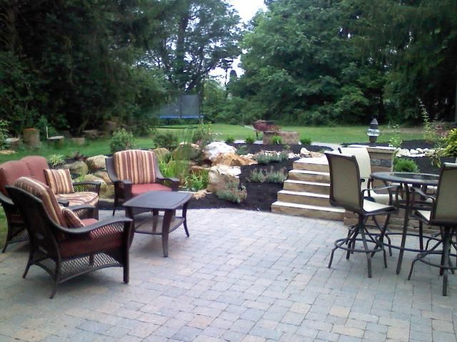 Patio with seating areas: wicker chairs with cushions, and a small table, near a stone structure and garden.