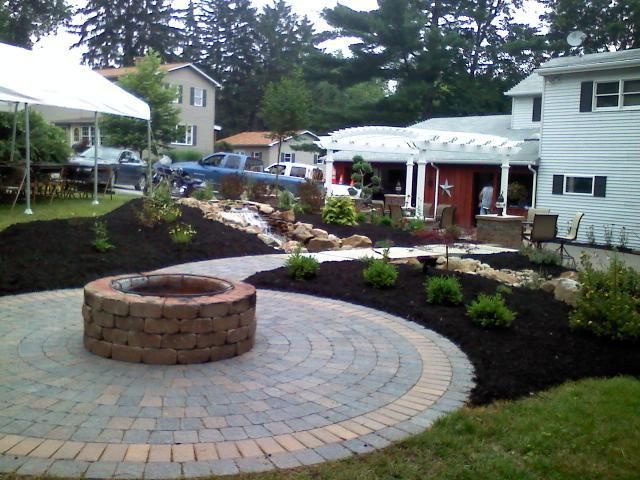 Circular brick patio with fire pit, garden bed, and house in background.