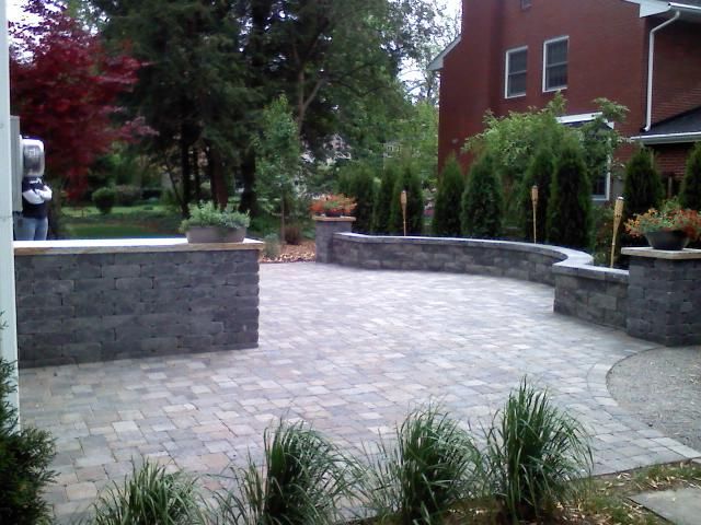 Brick patio with stone walls, surrounded by landscaping, beside a brick house.