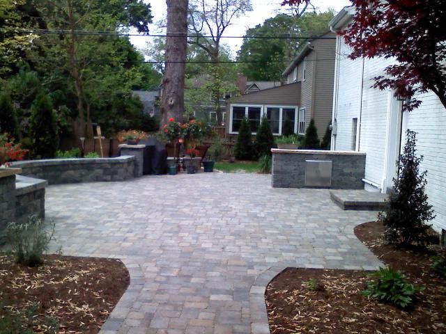 Paver patio with stone walls, grill, and landscaped yard. House in the background.