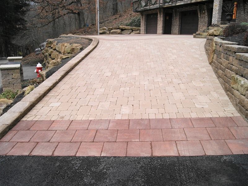 Brick paver driveway with dark red and tan bricks, leading to a house with a stone retaining wall.
