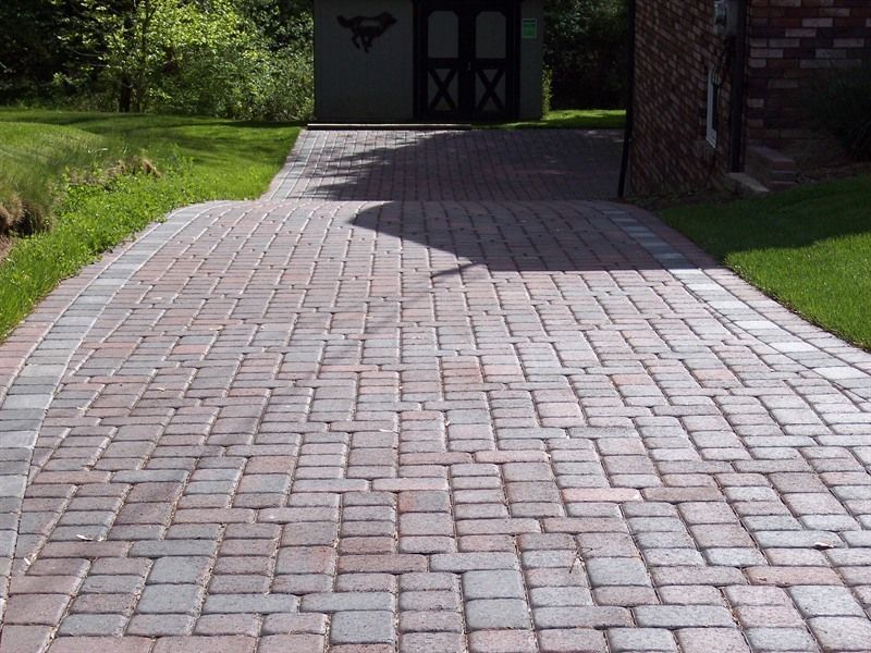 Brick driveway leading to a dark shed, flanked by grass and a brick building on a sunny day.