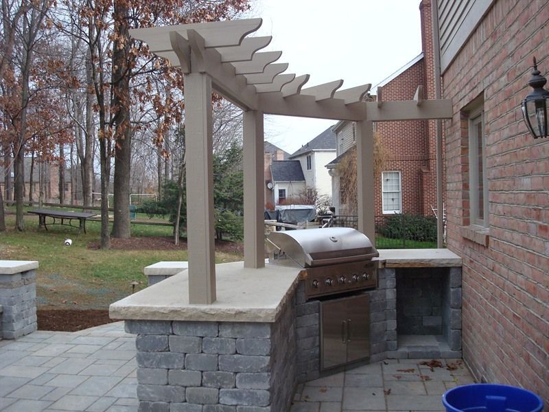 Outdoor kitchen with a grill under a pergola on a brick patio.