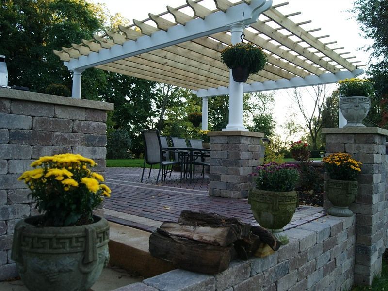 Pergola over brick patio, stone pillars with potted flowers, table and chairs.