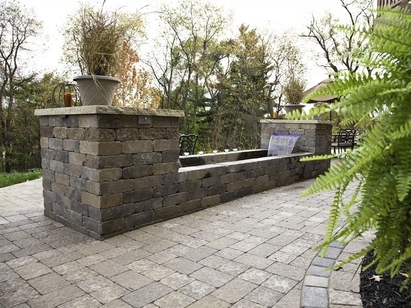 Stone water feature with brick patio, a potted plant, and greenery in the background.