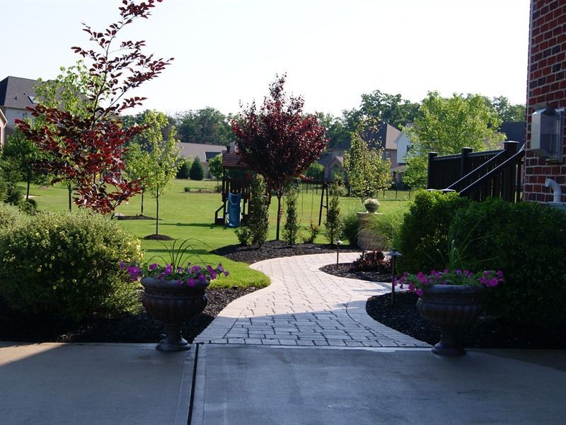 Brick path curves through a backyard, flanked by shrubs and trees, leading to a lawn and trees.
