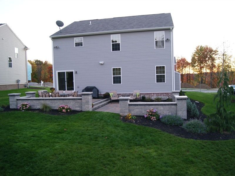 Rear view of a light gray house with a stone patio, retaining walls, and green lawn.