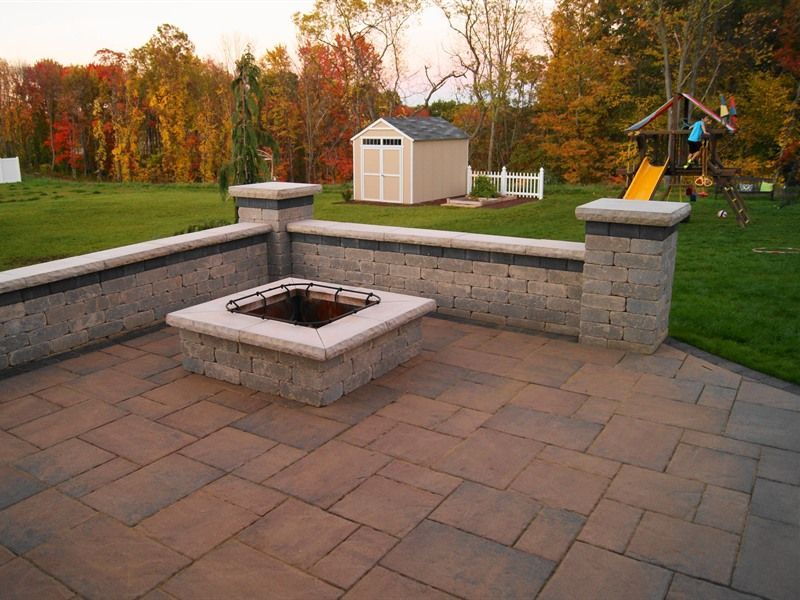 Fire pit area with brick patio, low wall, shed, and playground in a backyard with autumn foliage.