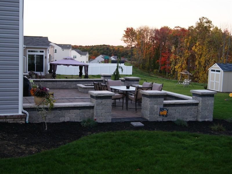Outdoor patio with a table, chairs, and built-in planters on a brick surface; trees in the background.