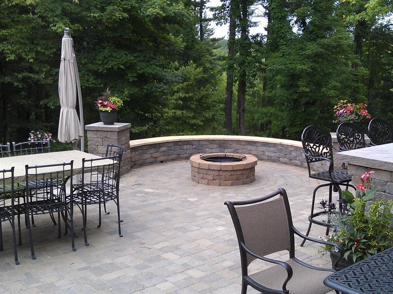 Patio with fire pit, stone wall, tables, chairs, and umbrella in a wooded setting.