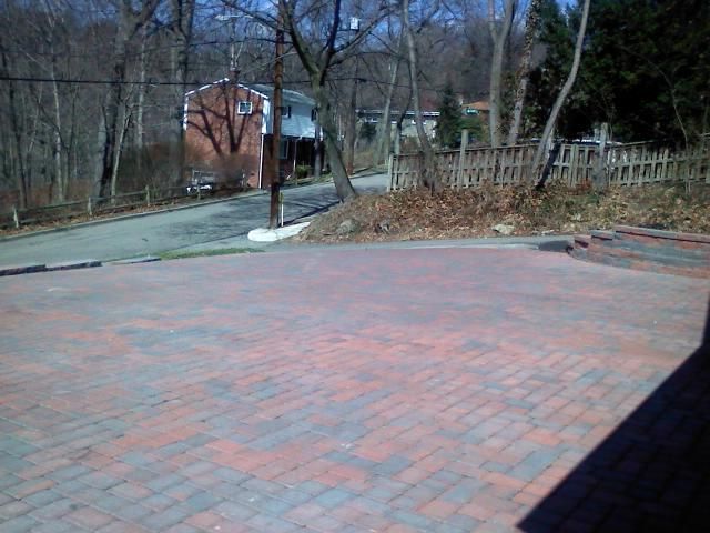 Brick paved driveway leading to a residential street with trees and houses.