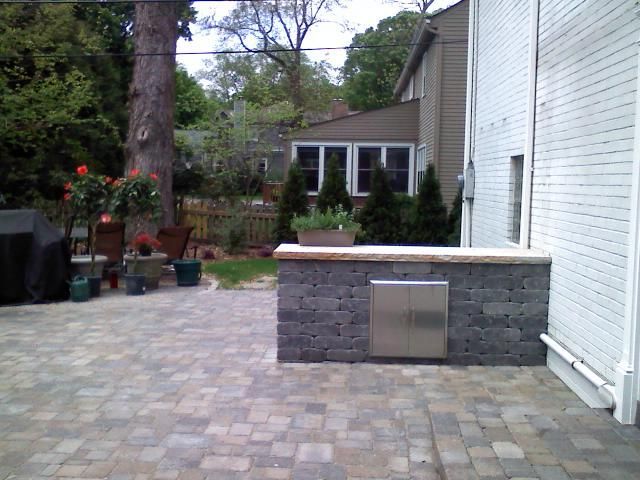 Backyard patio with brick pavers, built-in grill, and a house in the background.