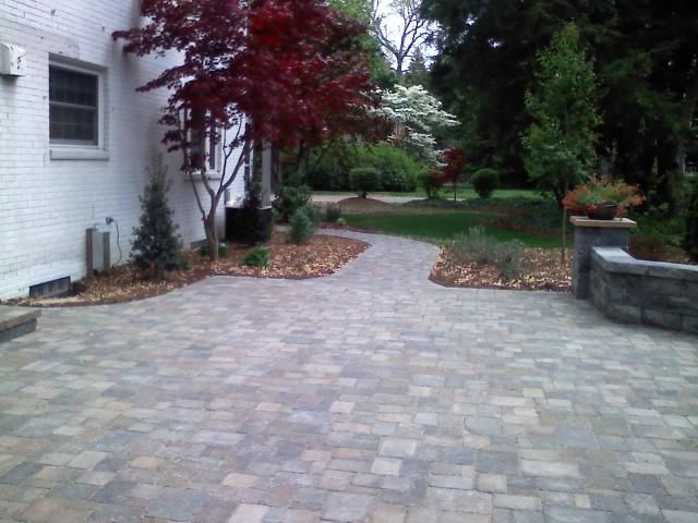 Paver patio with a winding path leading to a backyard garden, featuring trees and landscaping.