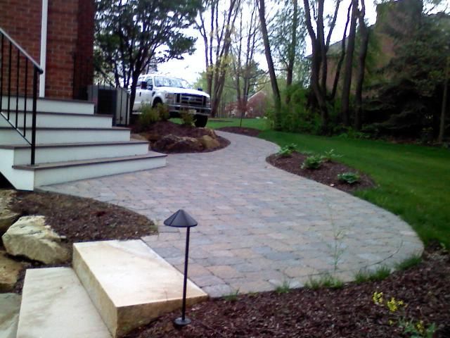 Stone pathway curving through a landscaped yard, leading to a white truck. Brick stairs on the left.
