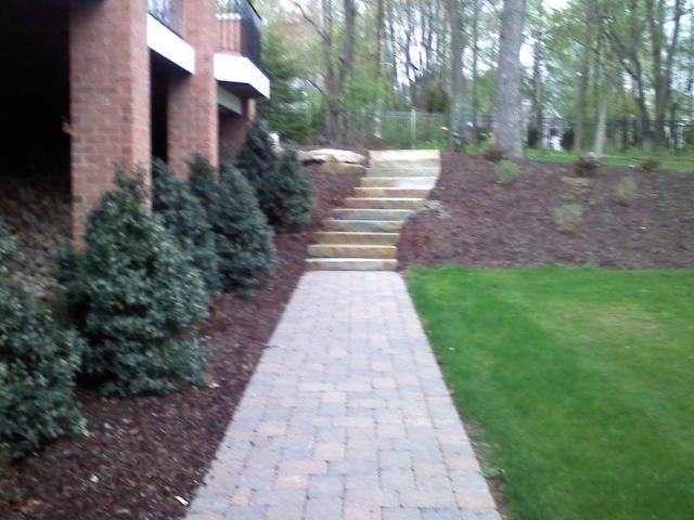 Brick walkway with steps and landscaping, leading uphill to a wooded area.