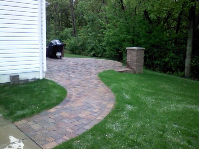 Brick paver pathway curves through a grassy yard next to a house; a grill and a pillar are visible.