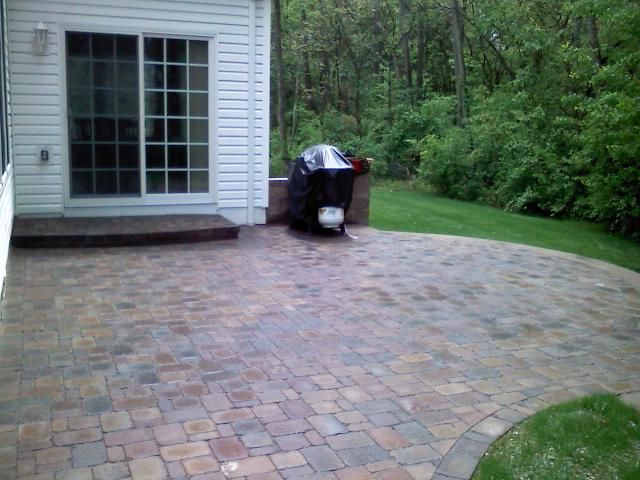 Brick patio next to a white house with a sliding door, grill, and lush green background.