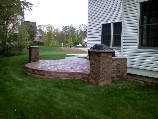 Stone patio with low retaining wall and pillars next to a house with white siding, surrounded by green grass.