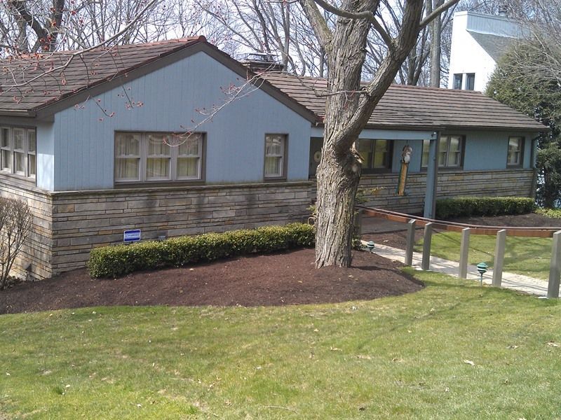 Blue house with brown roof, stone base, and green lawn. Brown mulch bed with hedge in front.