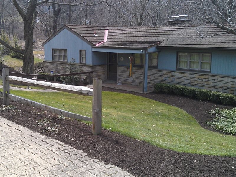 Blue house with stone facade, brown roof, and wooden fence. Green lawn and trees.