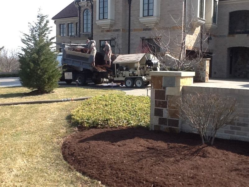 Landscapers unload mulch from a truck near a house, spreading it over a garden bed.