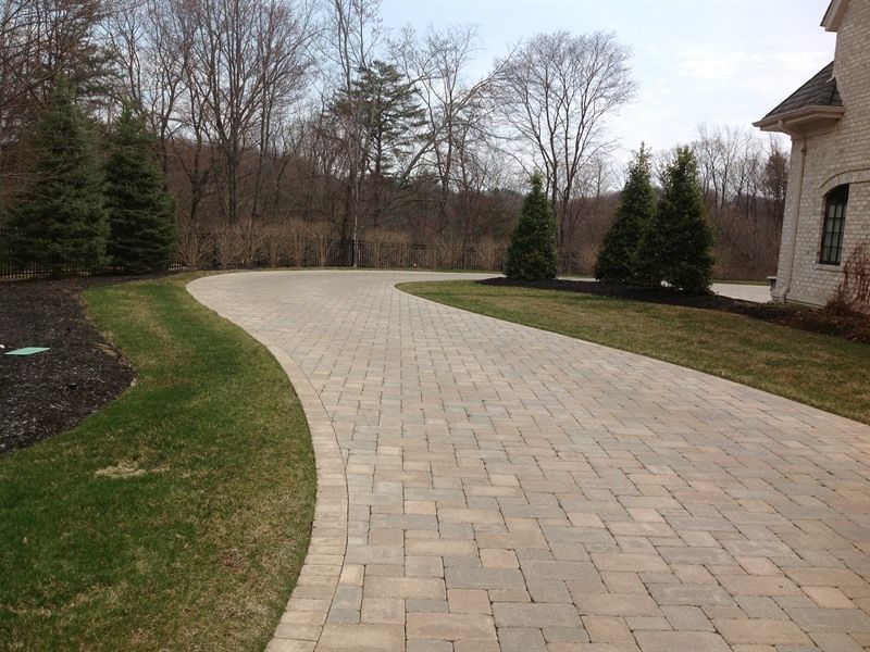 Brick driveway curving through a grassy yard toward a house, with trees in the background.